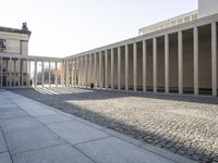 people walk past columns of a large building in a courtyard of stone and brick,