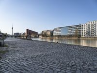 a large stone road with buildings and people riding bikes next to a river and two tall buildings