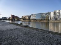 a large stone road with buildings and people riding bikes next to a river and two tall buildings