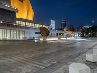 an empty courtyard with a stone bench outside of it, at night with buildings in the background