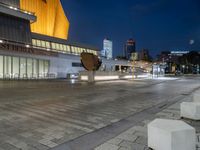 an empty courtyard with a stone bench outside of it, at night with buildings in the background