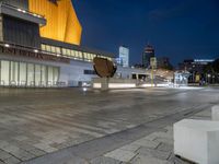 an empty courtyard with a stone bench outside of it, at night with buildings in the background