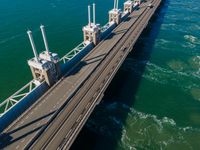 a bridge over the ocean with construction equipment in the background and two people walking on it