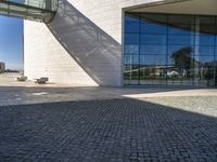 brick and glass building with benches sitting on the pavements in front of it and a sky scrape in the background