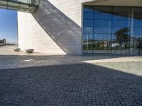 brick and glass building with benches sitting on the pavements in front of it and a sky scrape in the background