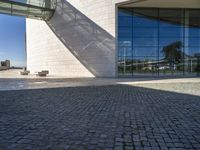 brick and glass building with benches sitting on the pavements in front of it and a sky scrape in the background