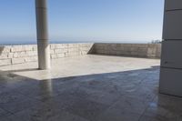 a patio with stone columns and flooring facing the ocean, a building in the background