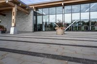 the walkway outside of the large building is stone and glass with several plants in flower pots on them