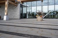 the walkway outside of the large building is stone and glass with several plants in flower pots on them