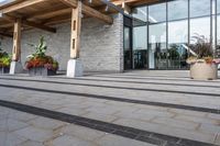 the walkway outside of the large building is stone and glass with several plants in flower pots on them