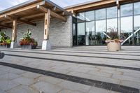 the walkway outside of the large building is stone and glass with several plants in flower pots on them