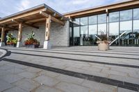 the walkway outside of the large building is stone and glass with several plants in flower pots on them