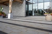 the walkway outside of the large building is stone and glass with several plants in flower pots on them