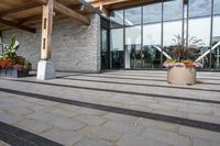 the walkway outside of the large building is stone and glass with several plants in flower pots on them