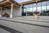 the walkway outside of the large building is stone and glass with several plants in flower pots on them