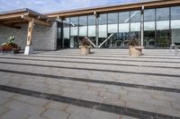 the walkway outside of the large building is stone and glass with several plants in flower pots on them