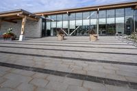 the walkway outside of the large building is stone and glass with several plants in flower pots on them