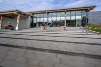 the walkway outside of the large building is stone and glass with several plants in flower pots on them