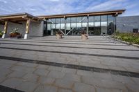 the walkway outside of the large building is stone and glass with several plants in flower pots on them