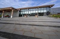 the walkway outside of the large building is stone and glass with several plants in flower pots on them