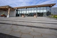 the walkway outside of the large building is stone and glass with several plants in flower pots on them