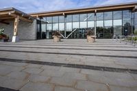 the walkway outside of the large building is stone and glass with several plants in flower pots on them