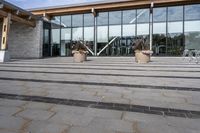 the walkway outside of the large building is stone and glass with several plants in flower pots on them