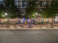 tables and benches sit under the trees at night on a sidewalk by a brick building