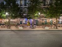 tables and benches sit under the trees at night on a sidewalk by a brick building