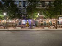 tables and benches sit under the trees at night on a sidewalk by a brick building