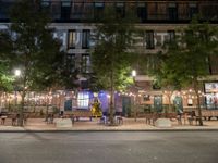 tables and benches sit under the trees at night on a sidewalk by a brick building