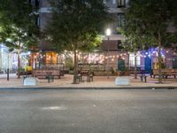 tables and benches sit under the trees at night on a sidewalk by a brick building