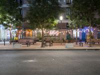 tables and benches sit under the trees at night on a sidewalk by a brick building