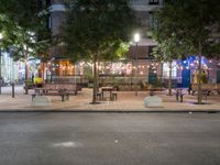 tables and benches sit under the trees at night on a sidewalk by a brick building