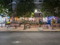 tables and benches sit under the trees at night on a sidewalk by a brick building