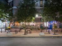 tables and benches sit under the trees at night on a sidewalk by a brick building