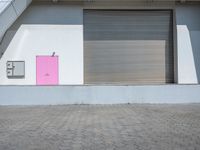 a bright pink door stands outside a white wall and brick floor of an open garage