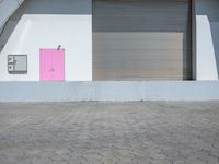 a bright pink door stands outside a white wall and brick floor of an open garage