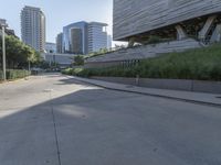 a paved, empty city street with grass along the sides and modern architecture on the hill