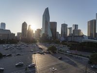 the sun is setting over a city with tall buildings and cars in a parking lot