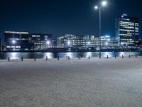 the lights are on at night beside the buildings and water below them, along with an empty brick sidewalk