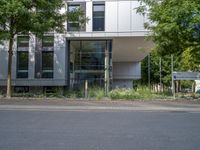 a large building with an entrance on a city street lined by tall green trees, and a small bench near it