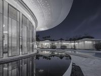 an outside view of a modern building at night with a pool area below that reflects the surrounding and beyond