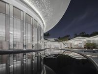 an outside view of a modern building at night with a pool area below that reflects the surrounding and beyond