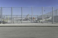 a gate with airplanes parked behind it next to a road and parking lot in the sun