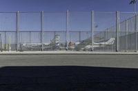 a gate with airplanes parked behind it next to a road and parking lot in the sun