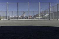 a gate with airplanes parked behind it next to a road and parking lot in the sun