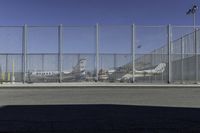 a gate with airplanes parked behind it next to a road and parking lot in the sun