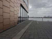 a large long building that has glass windows and a brick sidewalk near it on the side of a river