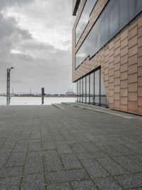 an empty sidewalk next to the water in front of a building with large glass windows